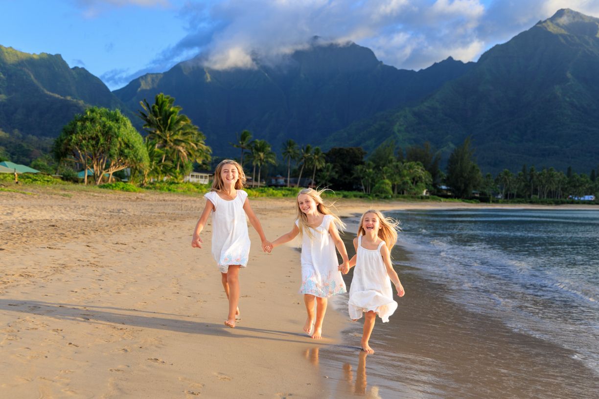 Three children in white dresses running along a beach with mountains in the background.