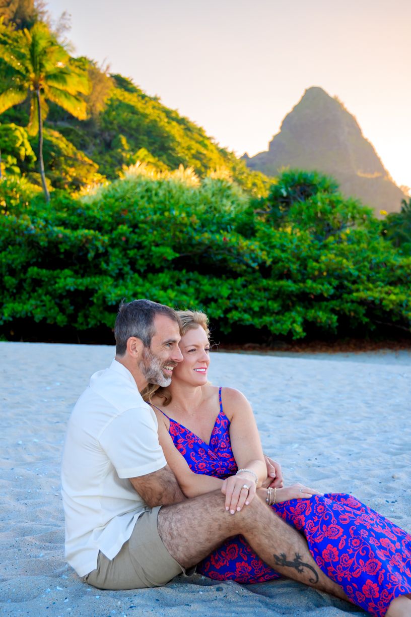 A couple relaxes on a tropical beach with mountains and palm trees during golden hour.