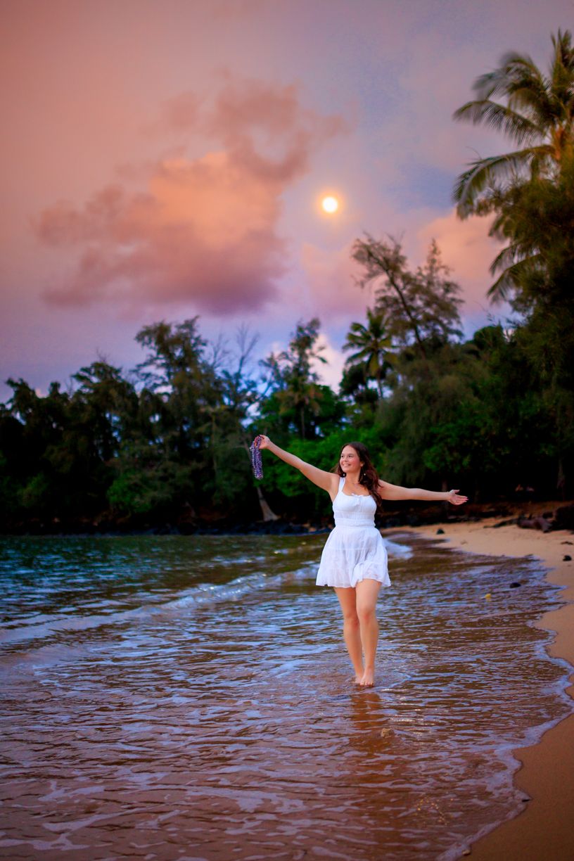 Woman in white dress standing in ocean water at sunset with palm trees in background.