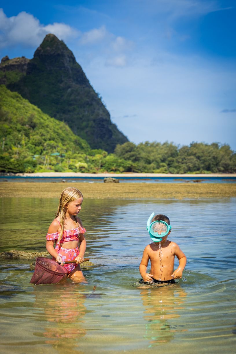 Two children playing in shallow water with a mountain peak backdrop in Hawaii.