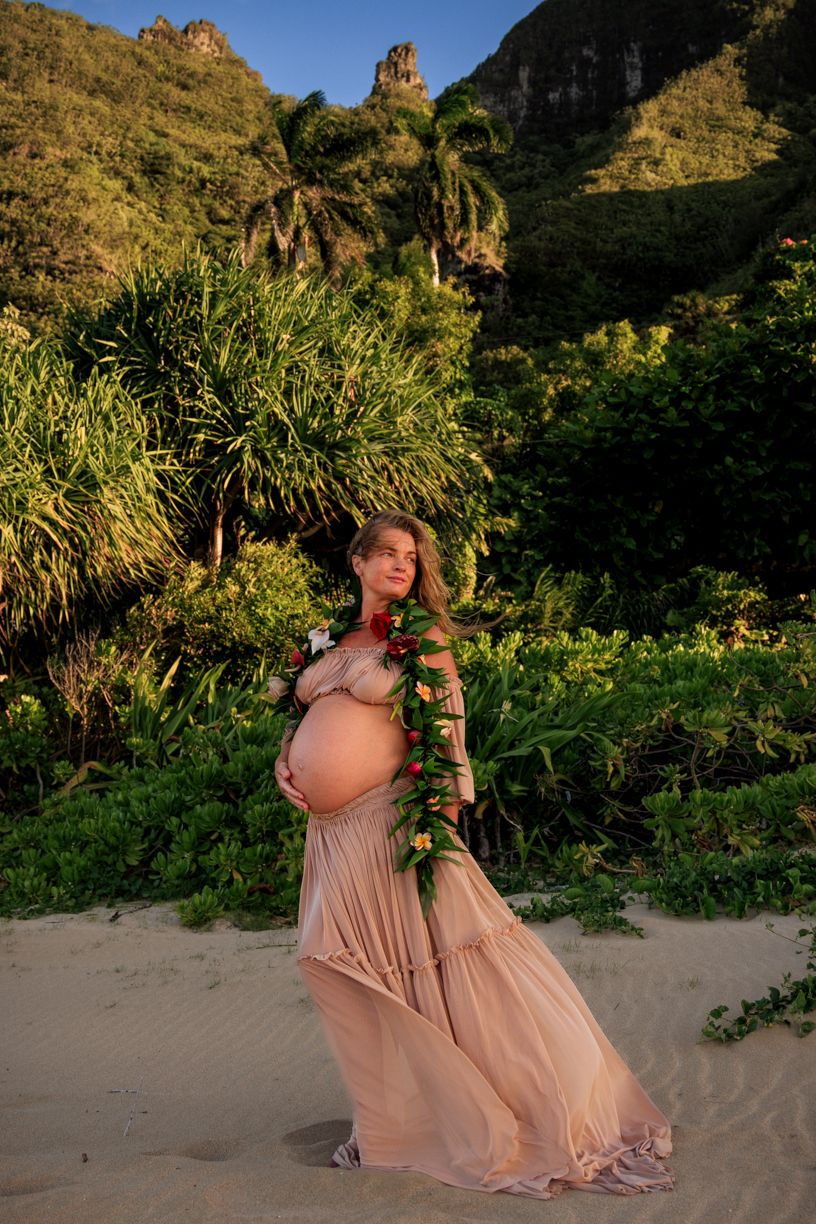 Expecting mother in flowing dress poses on beach with dramatic mountain landscape.
