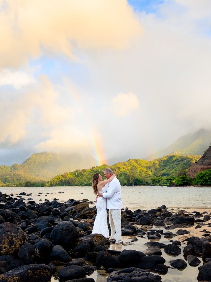 Couple embraces on black lava rocks with dramatic Hawaiian mountains and ocean in background at sunset.