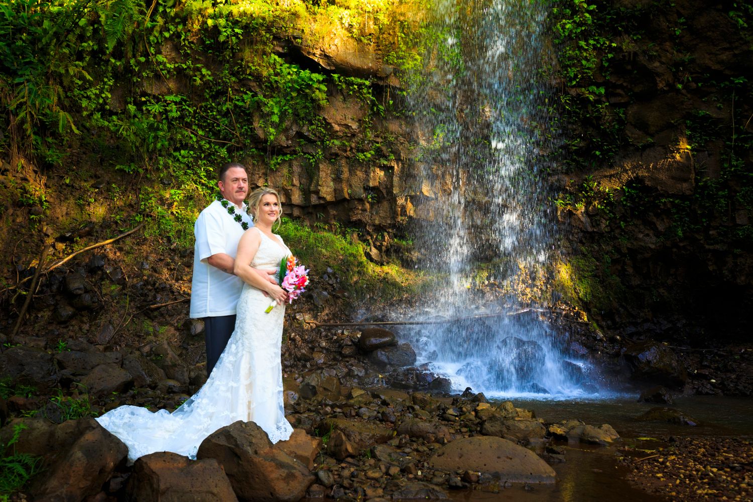 Wedding couple poses near scenic waterfall surrounded by tropical Hawaiian foliage.