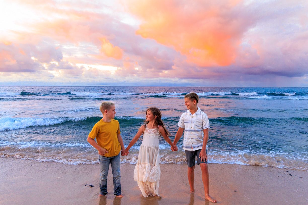 Three children hold hands on a sandy beach at sunset with colorful pink and blue clouds over ocean waves.
