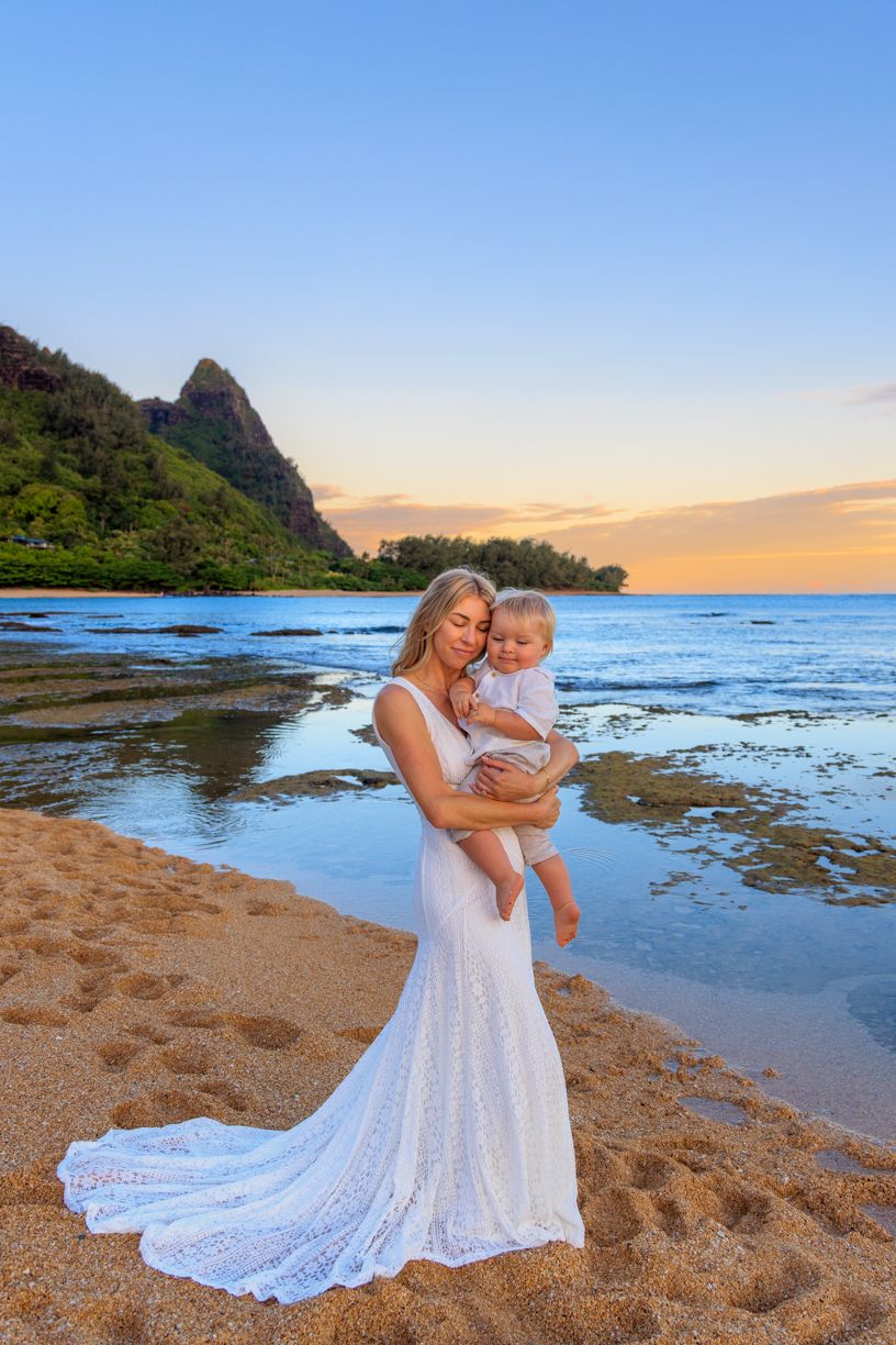 Mother and child in white dresses embracing on a scenic beach during golden hour at Tunnels Beach