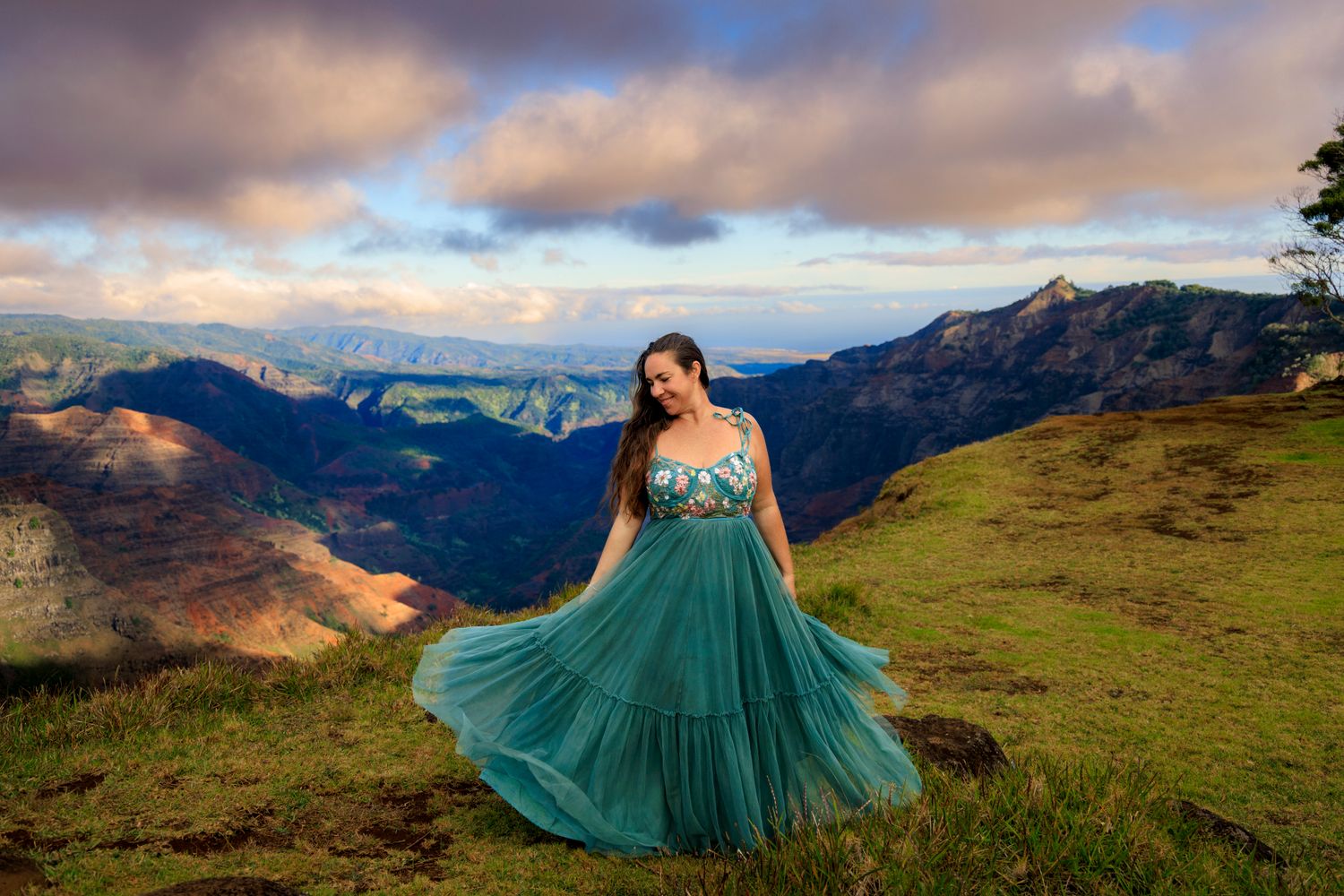 Woman in teal gown poses on green hillside with dramatic mountain valley and colorful sunset sky backdrop.