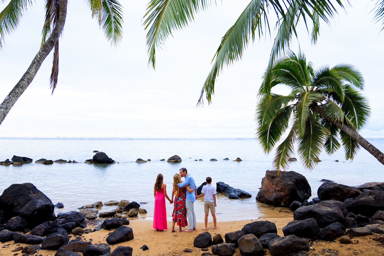 Family of three standing on a tropical beach with palm trees and black rocks in Hawaii.