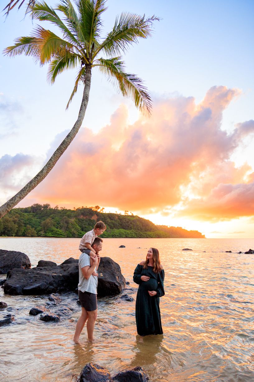 Pregnant woman in black dress with family at tropical beach during golden sunset, palm tree and colorful sky backdrop.