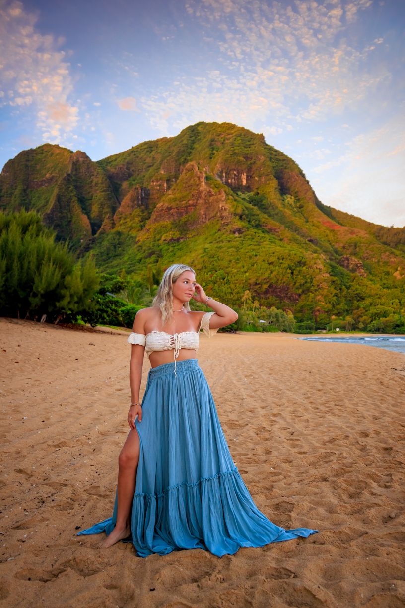 Beach photoshoot featuring blue dress against dramatic mountain landscape at golden hour.