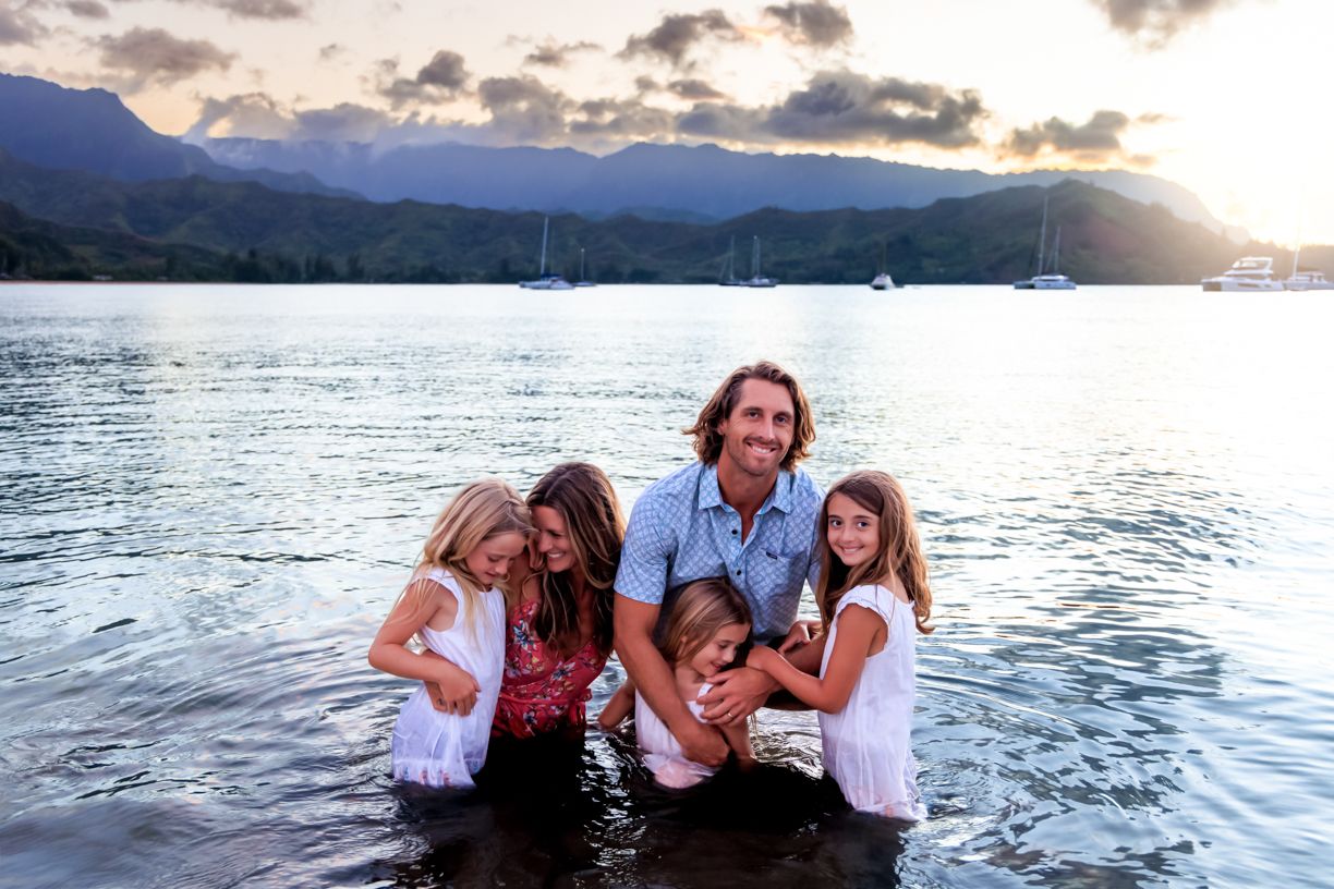 A family enjoying the waters of Hawaii at sunset with mountains and boats in the background.