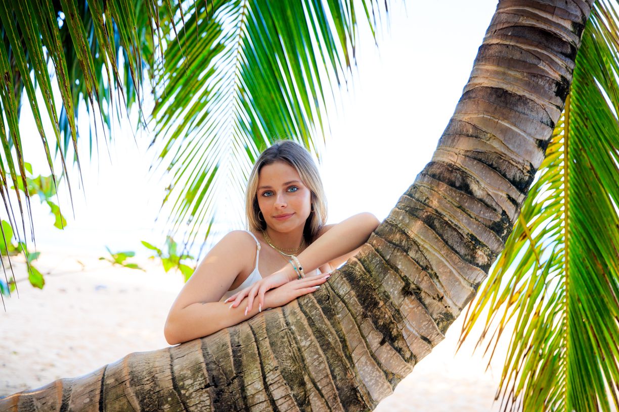 Woman leaning on palm tree trunk on tropical beach with sand and ocean in background.