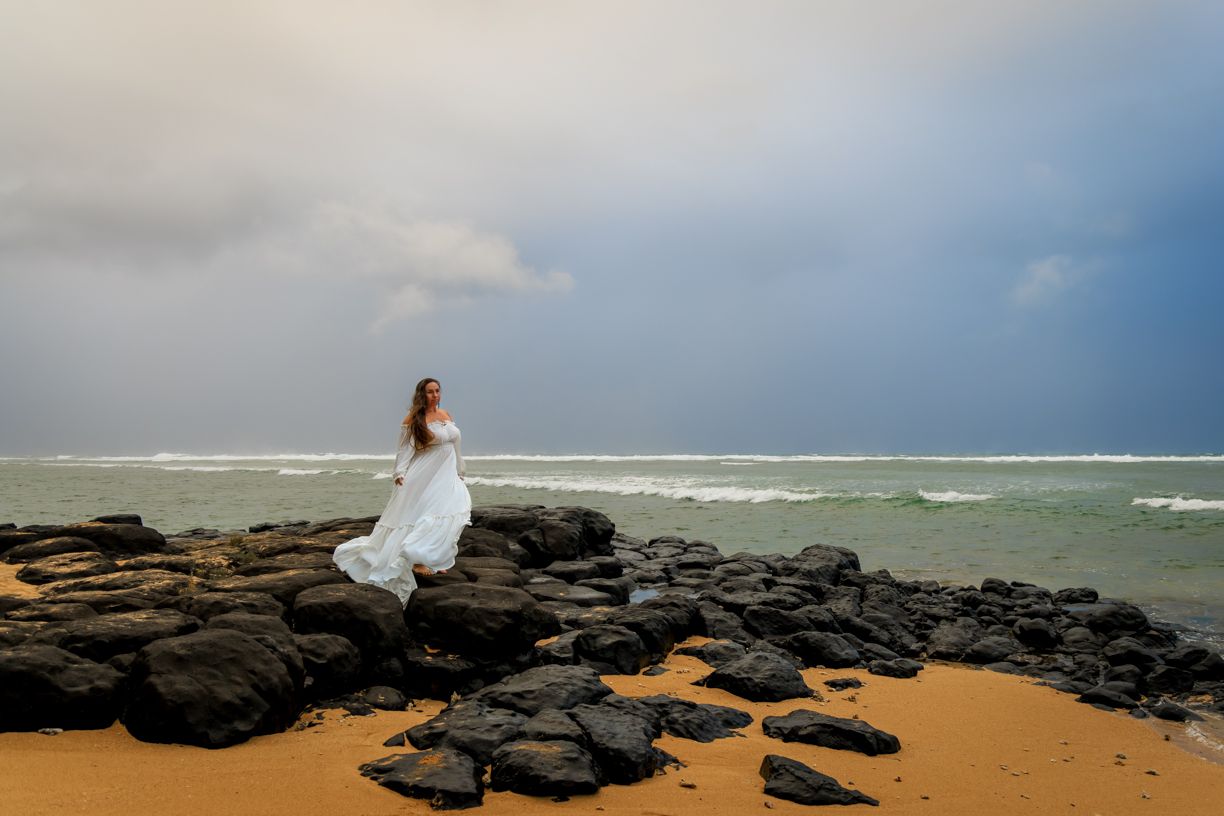 A white dress contrasts against dark volcanic rocks on a stormy beach.