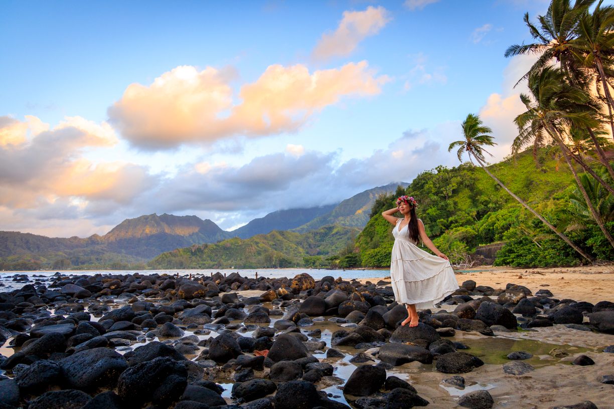 Palm trees line a Hawaiian beach at sunset with volcanic rocks and mountains in the distance.