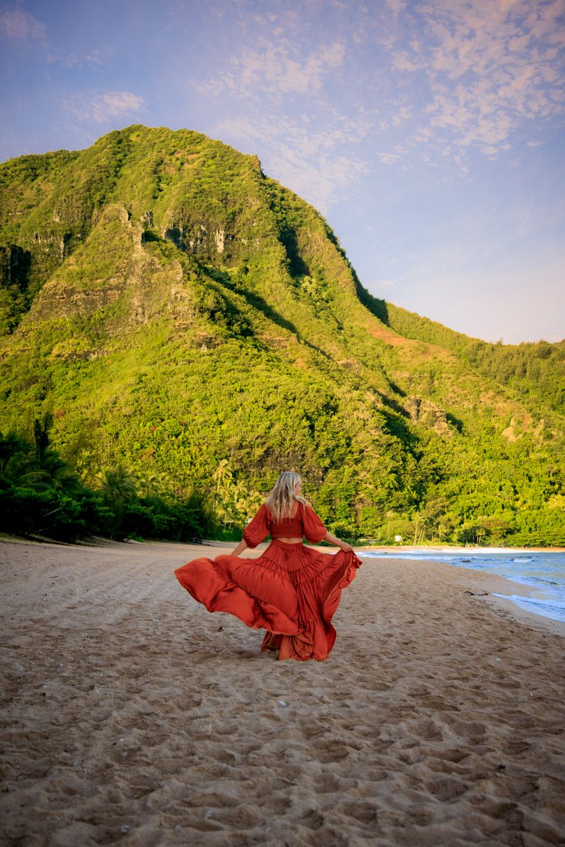 Red dress twirls dramatically on beach with towering green mountains in background.