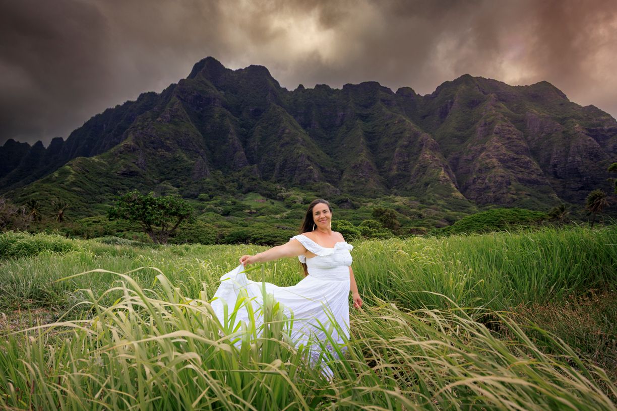 Woman in white dress standing in lush green field with dramatic mountain backdrop under stormy sky in Hawaii.
