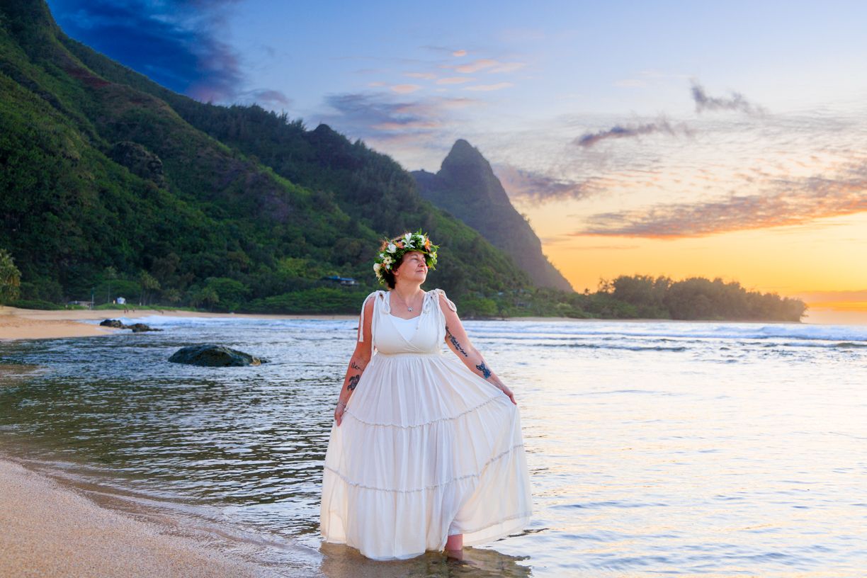 Woman in white dress and floral crown stands on Hawaiian beach at sunset with dramatic mountain backdrop.