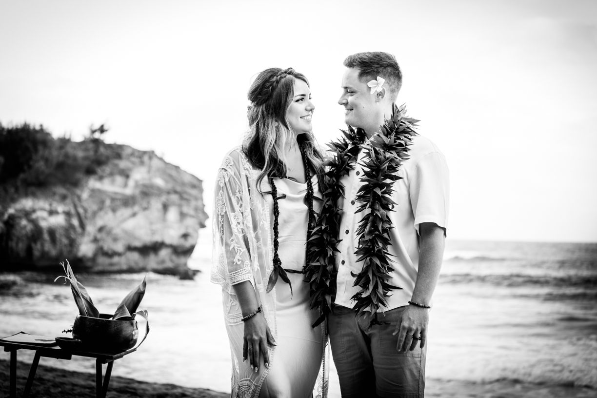 A black and white beach photo of a couple wearing traditional Hawaiian leis against a rocky coastal backdrop.