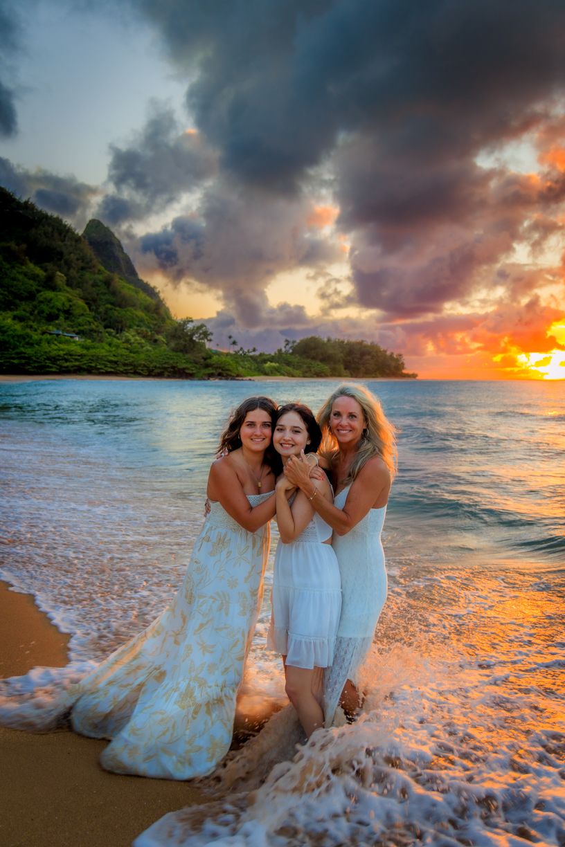 Three people in light dresses embrace on a beach at sunset with dramatic mountain and ocean backdrop.