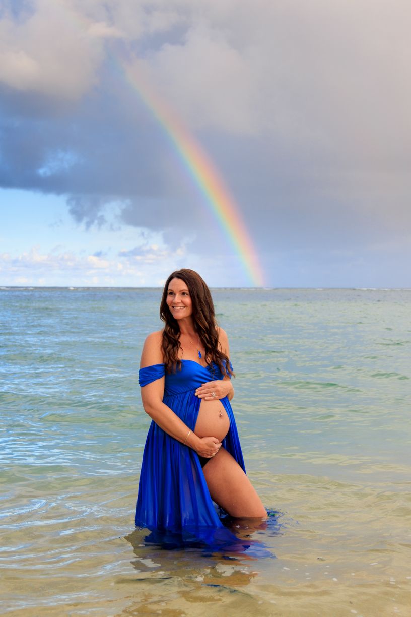 Maternity water session with rainbow backdrop and flowing blue dress.