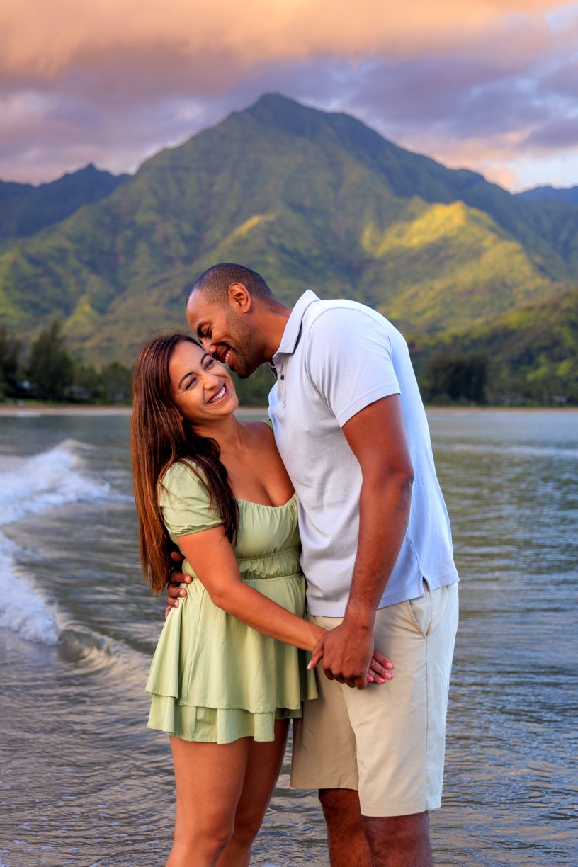 Romantic couple poses together at sunset on a Hawaiian beach with dramatic mountains in the background.