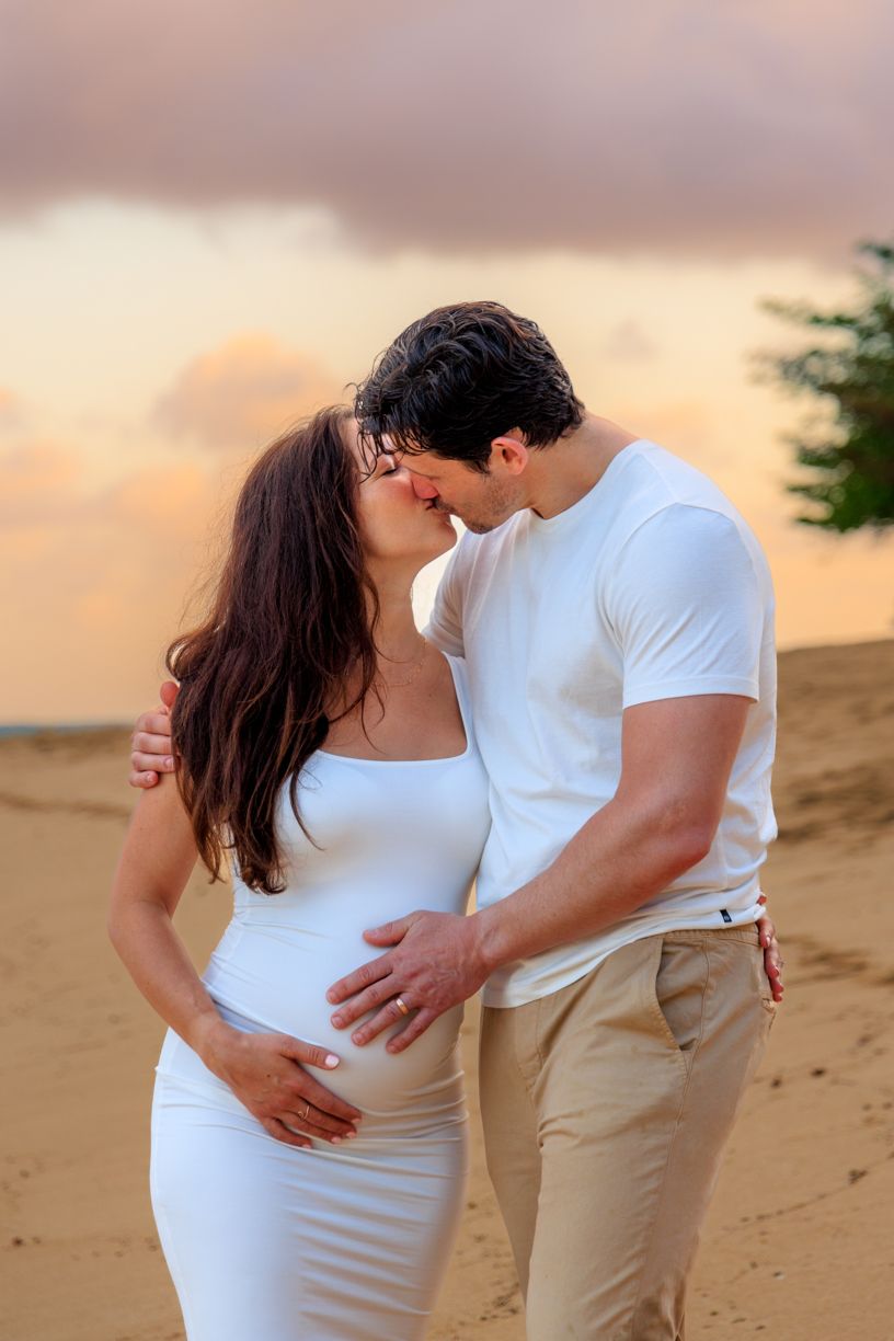 A maternity photo session on a sandy beach during sunset shows a couple in white and khaki.