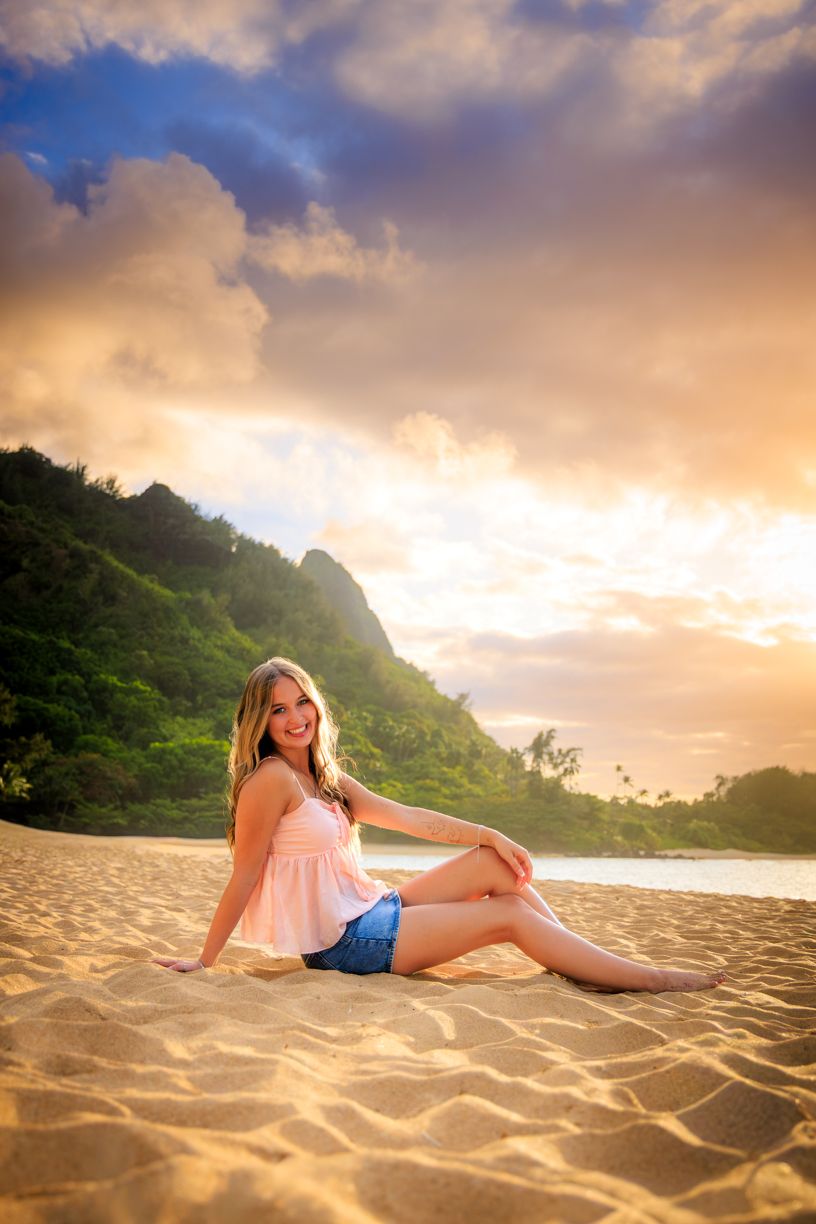 Woman in pink top and shorts sitting on sandy beach at sunset with mountains in background.