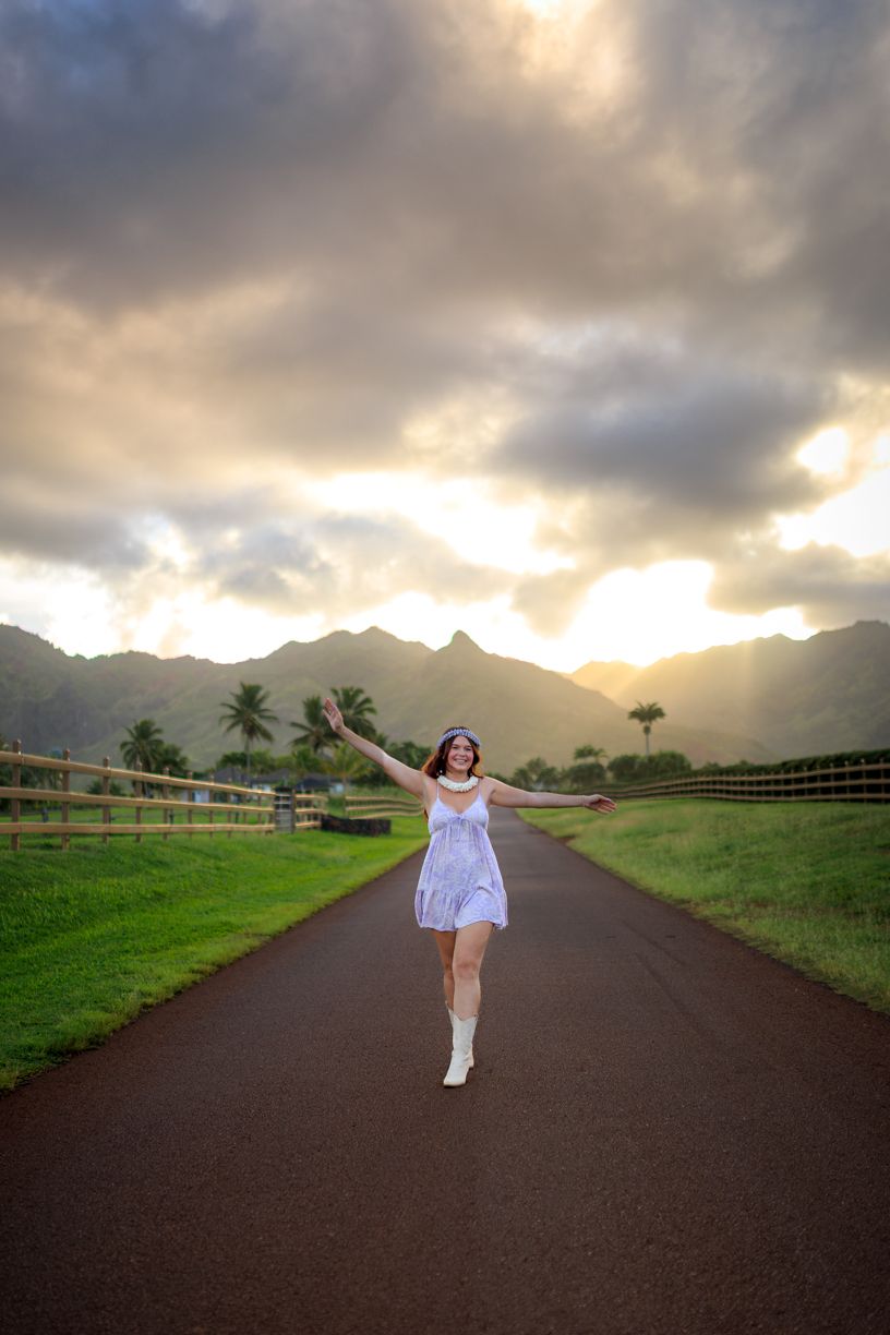 Person in white dress twirls on a country road surrounded by green fields and mountain views at dusk.