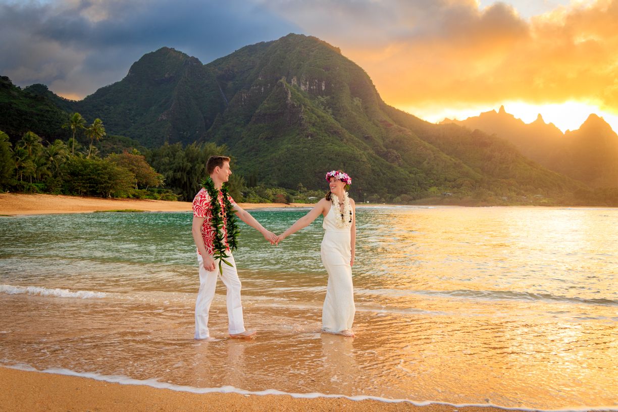 Couple in wedding attire holding hands on beach at sunset with mountain backdrop.