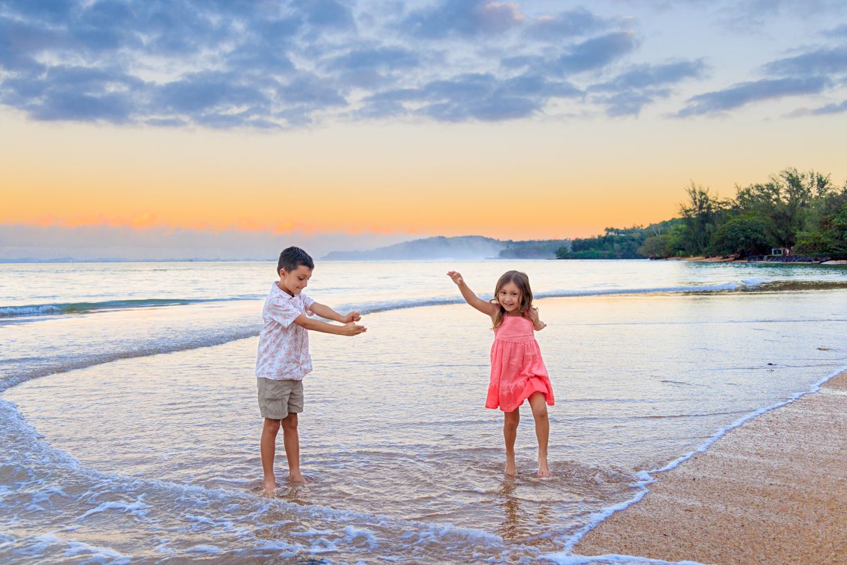 Two children playing in ocean waves on a sandy beach at sunset with colorful sky and distant mountains.