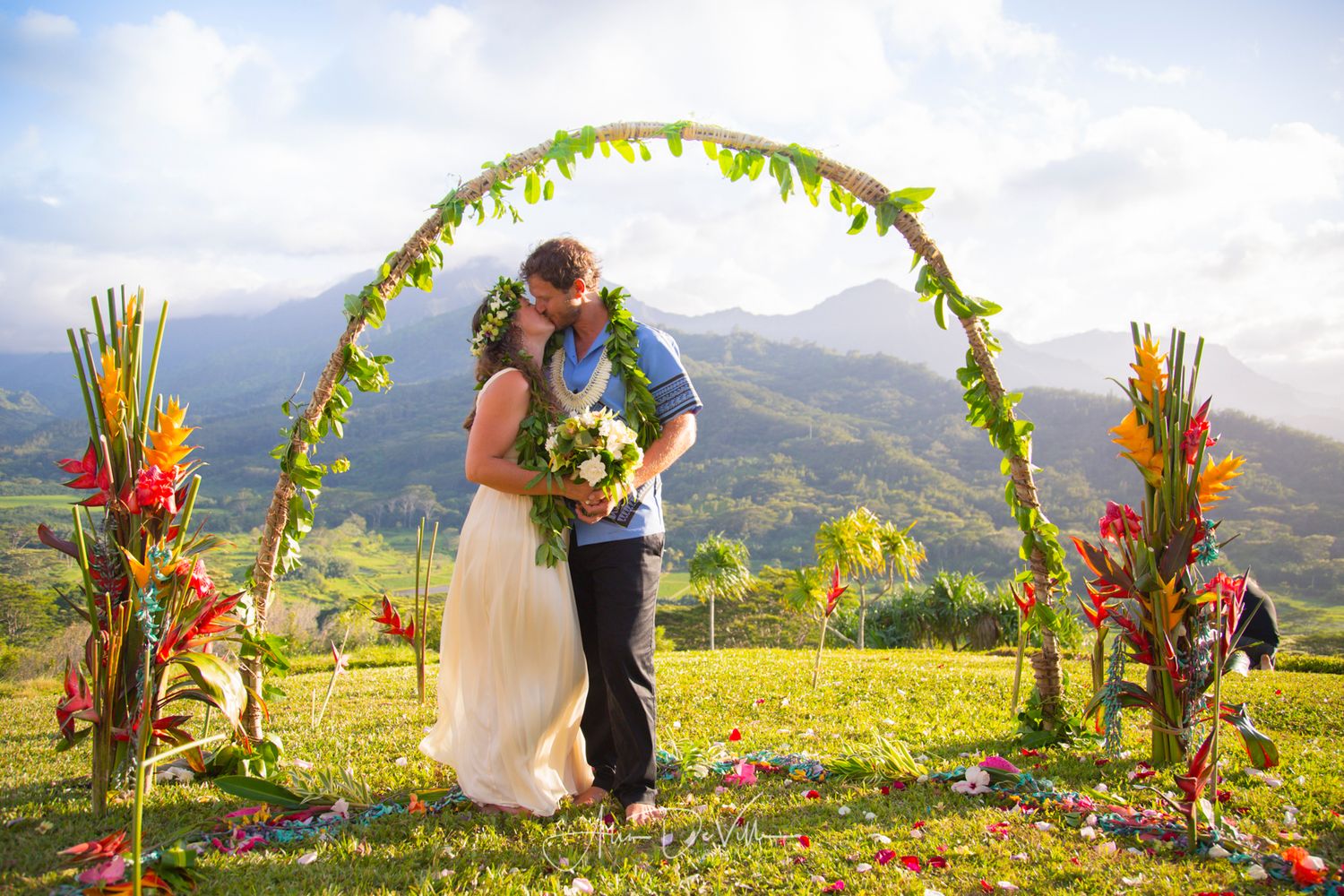 Tropical wedding ceremony under a yellow and red floral arch with mountain backdrop.