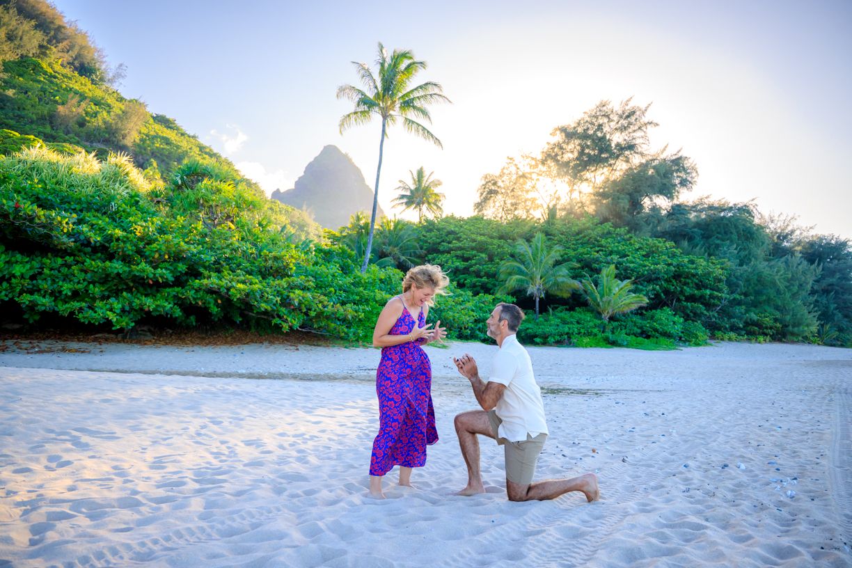 A beach proposal scene with mountains and palm trees in the background.