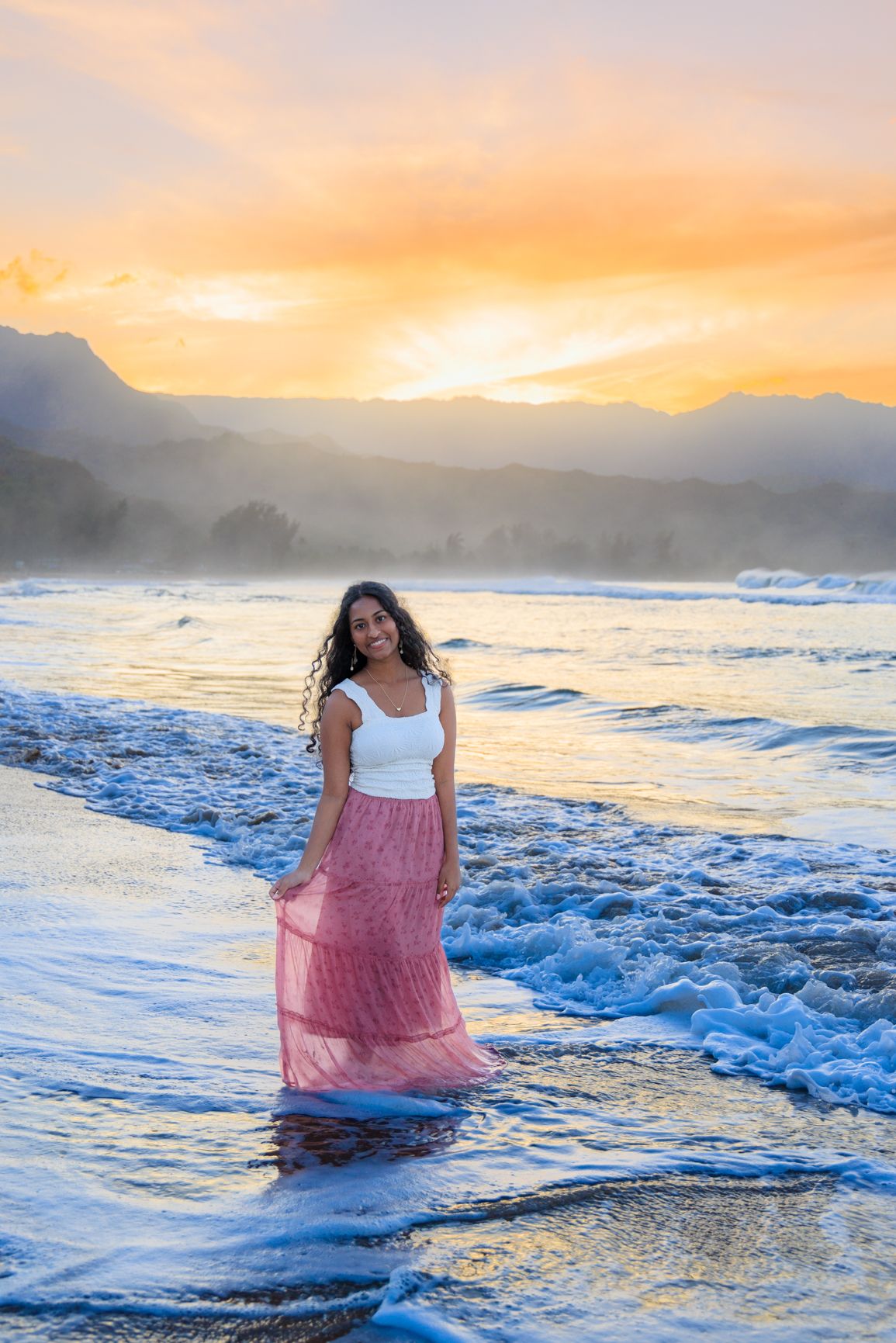 Woman in white top and pink skirt standing in ocean waves at sunset with mountains in background.