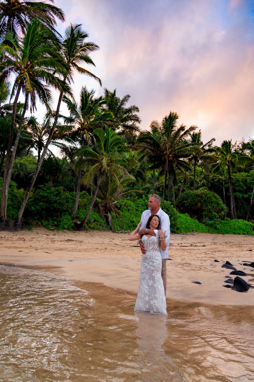 Couple in wedding attire standing in shallow water on tropical beach with palm trees.