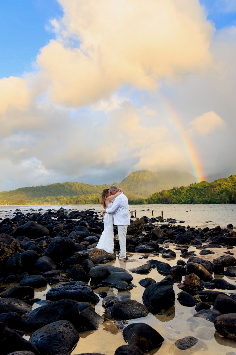 Scenic Hawaiian beach ceremony with black lava rocks and a rainbow appearing in the cloudy sky.