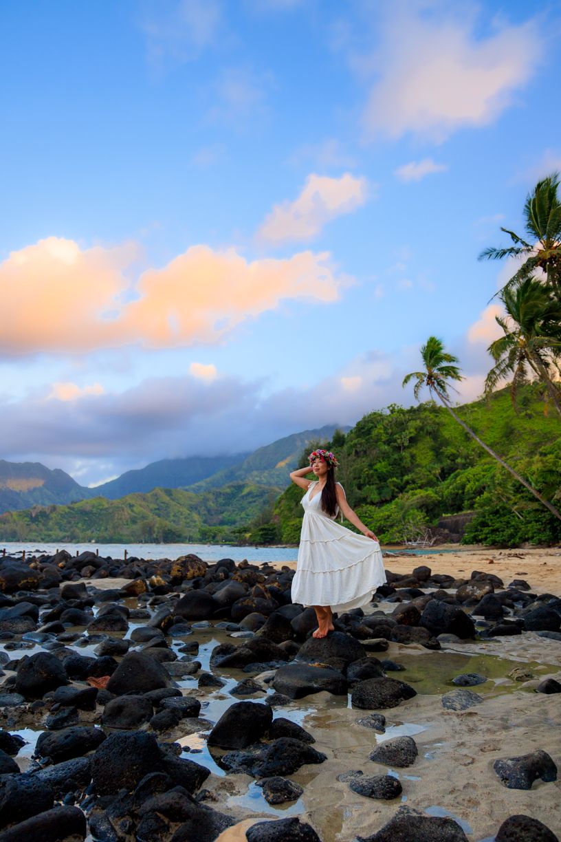 Woman in white dress on rocky beach with palm trees and mountains.