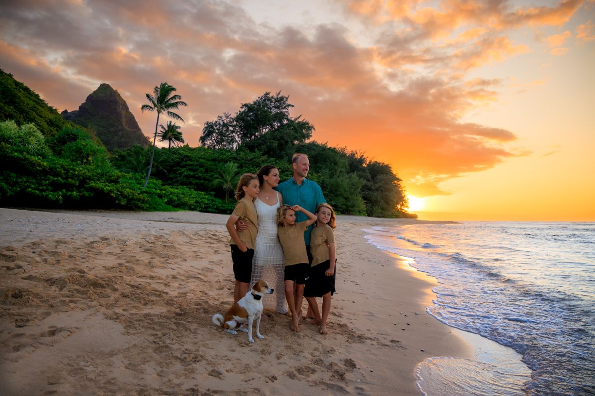 Family enjoying sunset moments on a Hawaiian beach with mountains and colorful sky in background.