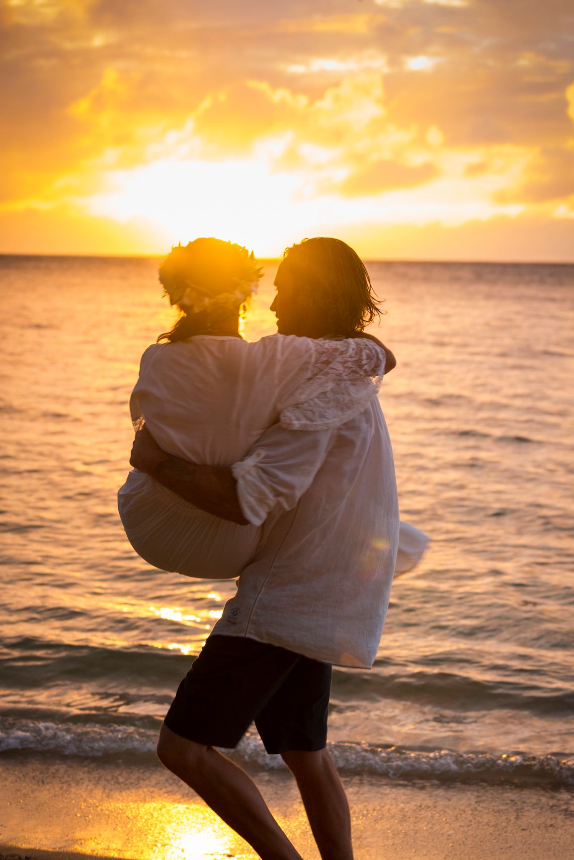 Silhouetted couple embracing on beach during golden Hawaiian sunset.