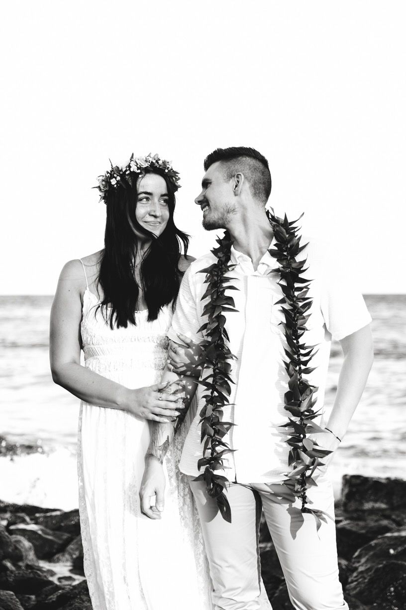 Intimate beach portrait session with Hawaiian leis and white attire against ocean waves.