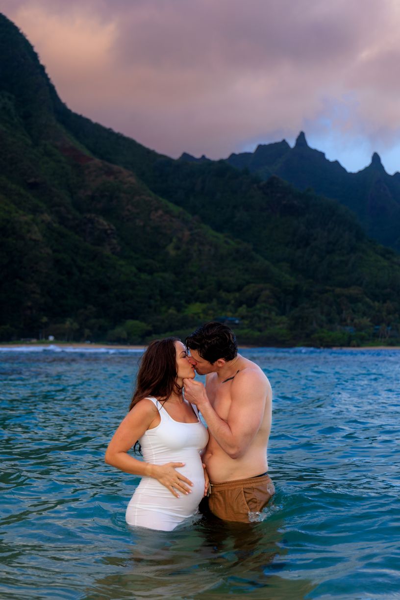 Couple embracing in tropical ocean waters with dramatic mountain landscape in the background at sunset.