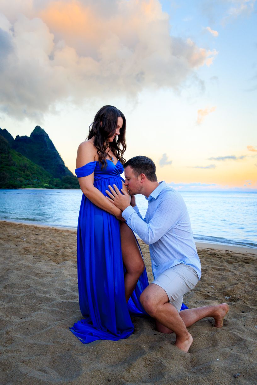 A romantic couple in blue and white outfits share an intimate moment on a beach at sunset with mountains in the background.