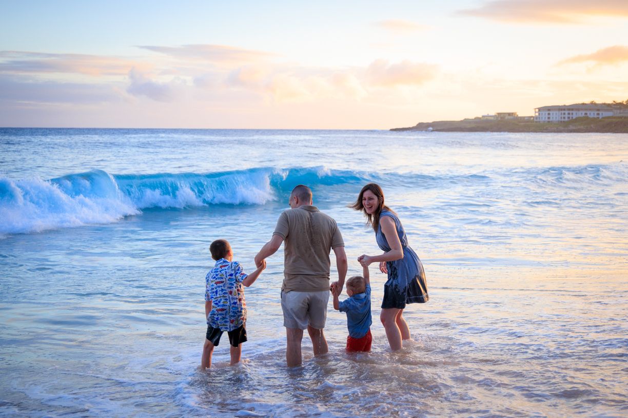Family playing in ocean waves during sunset with mountains in background.