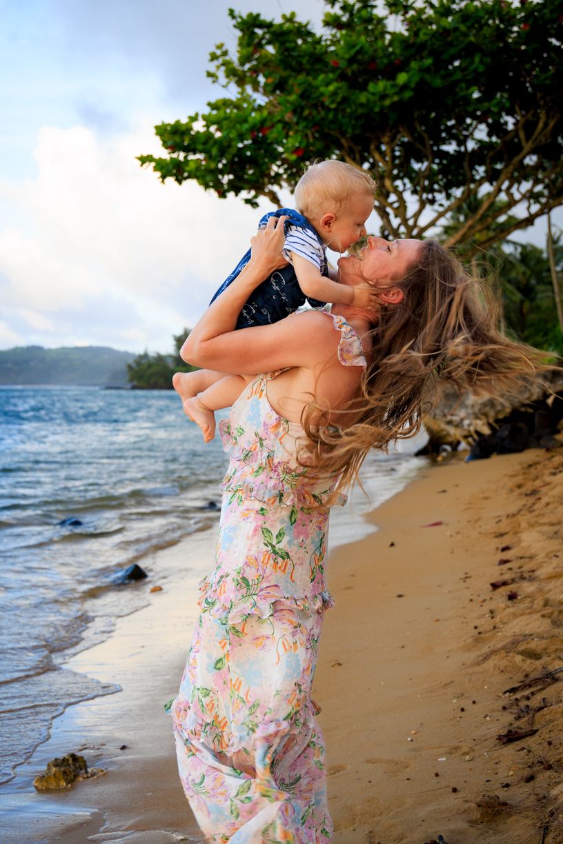 Woman in floral dress lifting baby on beach with ocean and trees in background.
