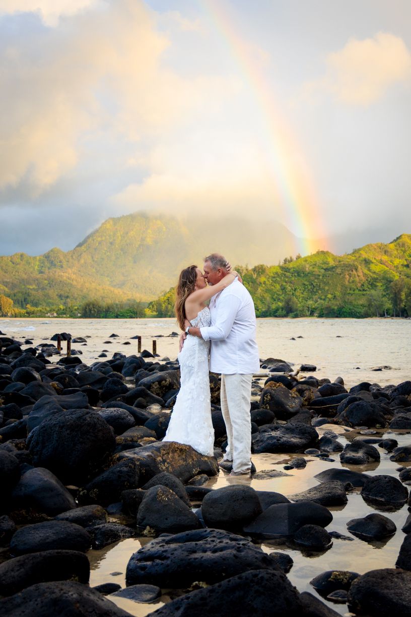 Newlyweds kissing on rocky beach with rainbow and mountain in background.