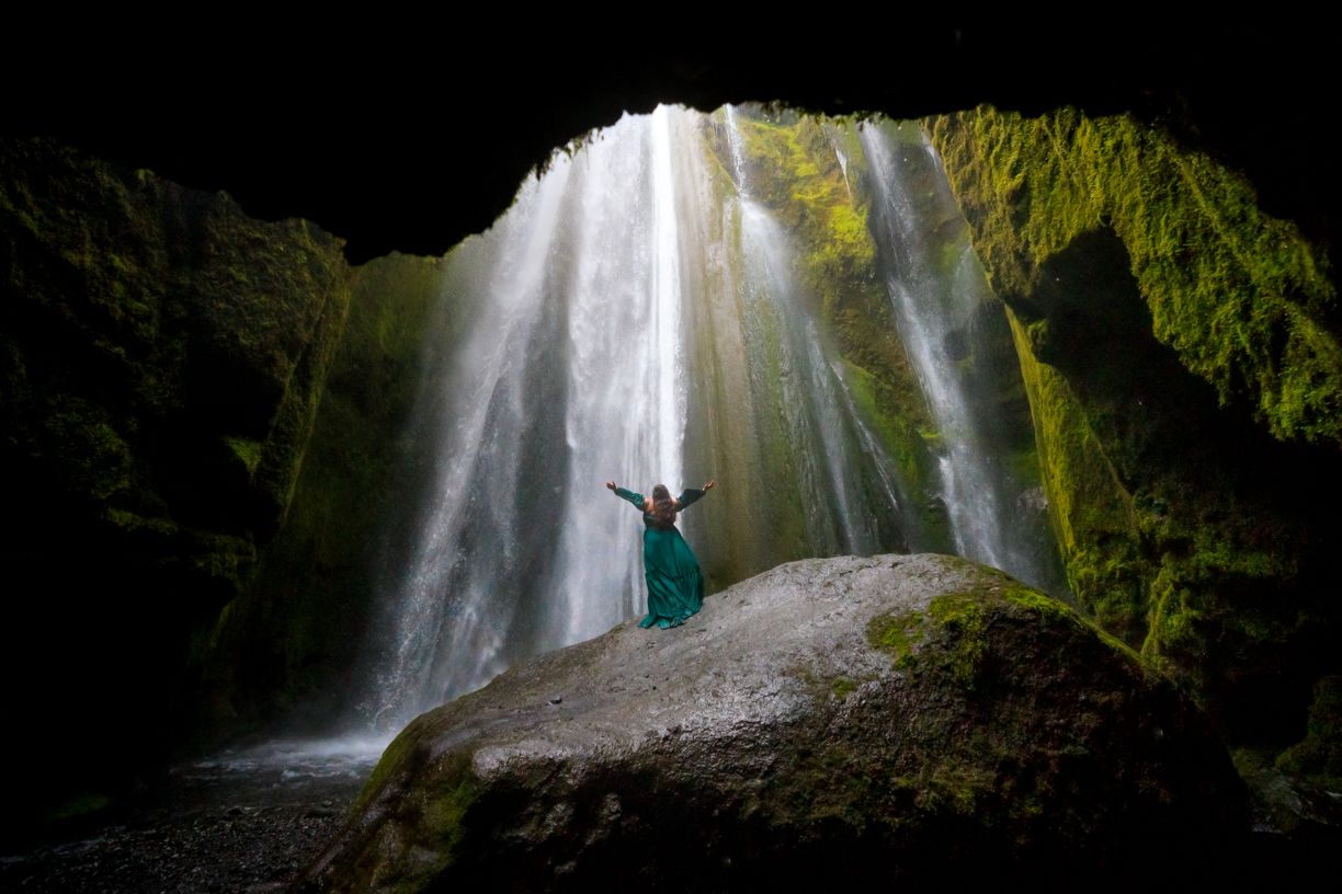 Dramatic waterfall cascades through dark cave opening with figure silhouetted against the light.
