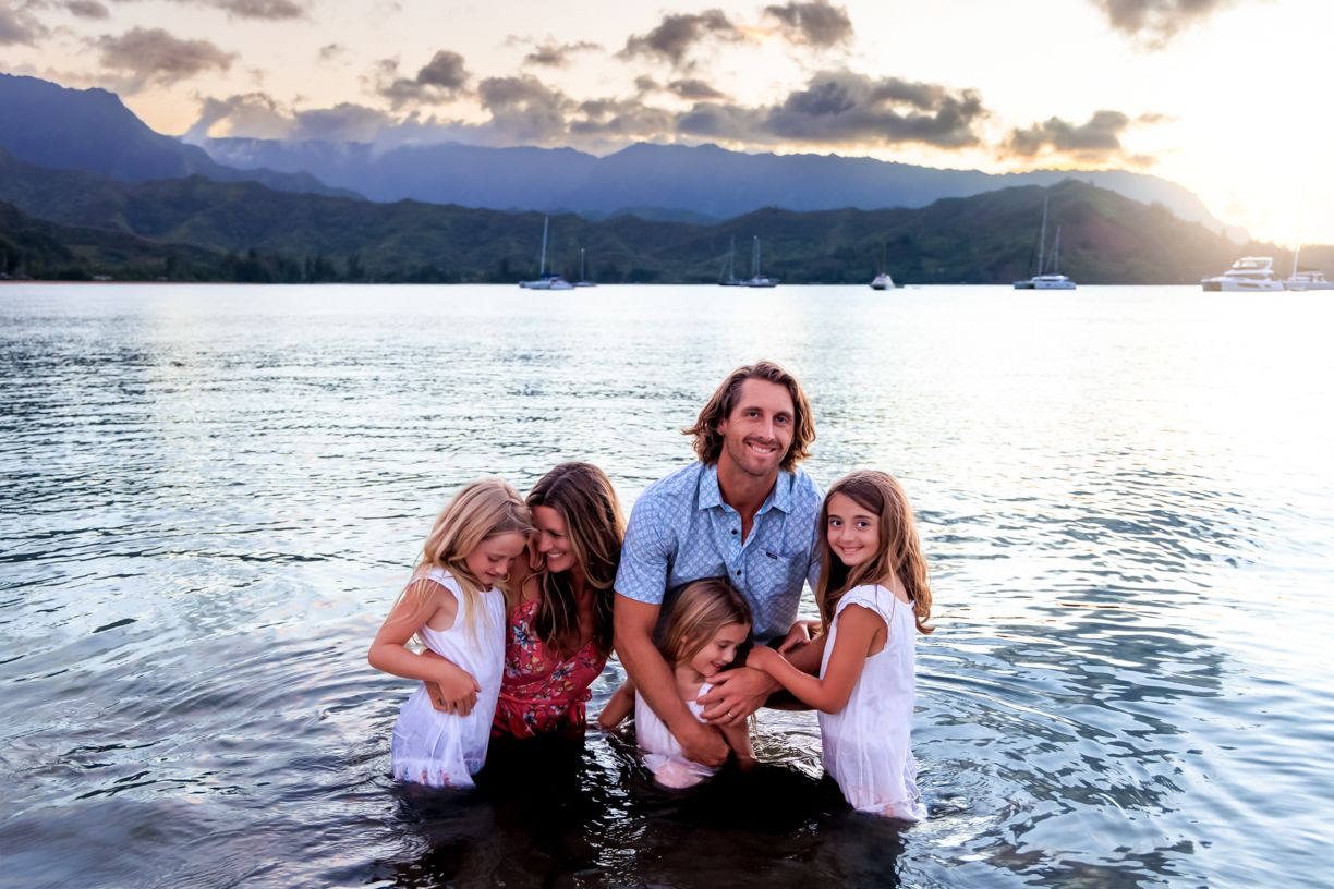 Family bonding moment in ocean water at sunset with mountains and boats in background.