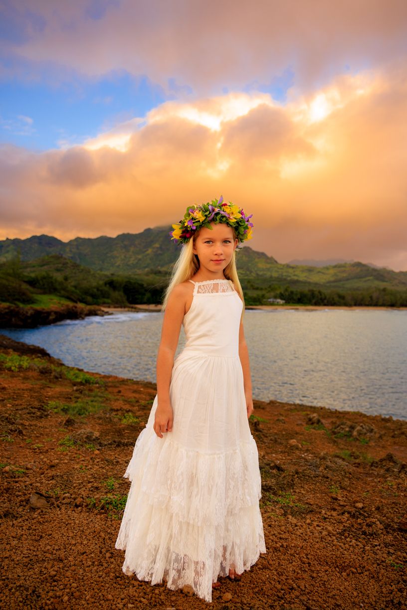 White lace dress and flower crown photographed at sunset by ocean with mountains in background.