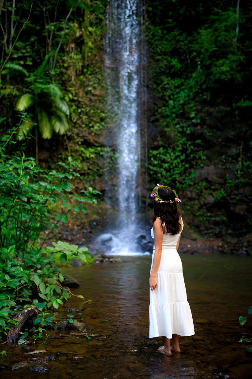 Two people in white dresses pose near a tropical waterfall in lush Hawaiian forest.