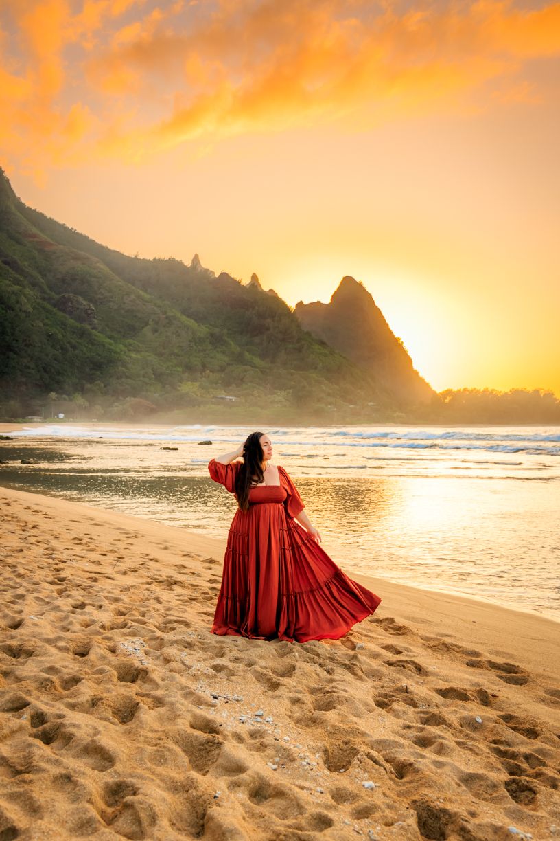 A flowing red dress twirls on a sandy beach at sunset with jagged mountains in the background.