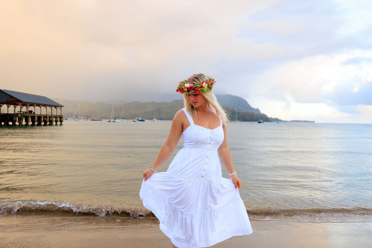Woman in white dress holding skirt on beach with pier and mountains in background.