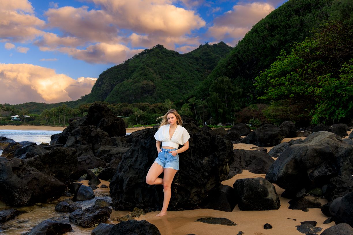 Person in white shirt standing on rocky beach with mountains and ocean in background.